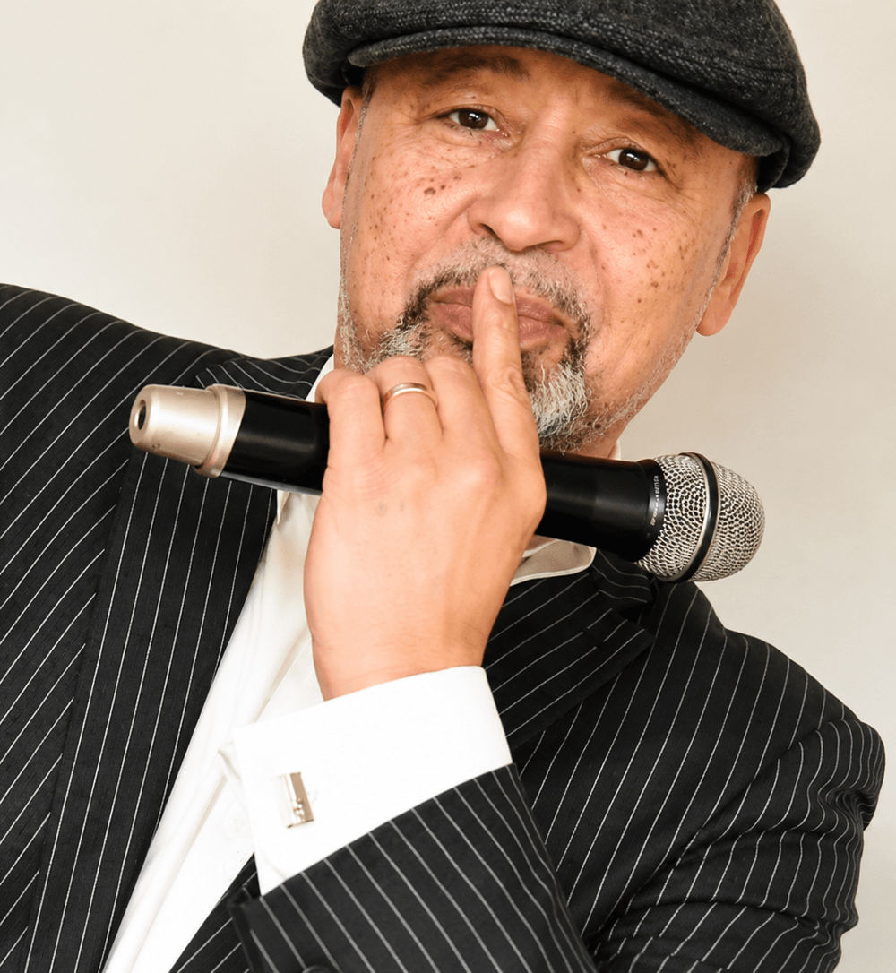 Geno Eccles wearing a black pinstripe suit and flat cap, holding a microphone and making a shushing gesture.